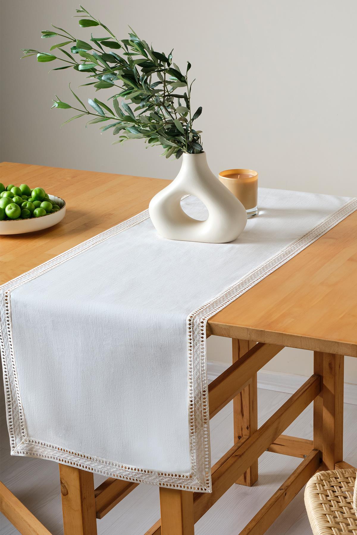 Wooden table decorated with a lace runner, featuring a modern vase and green plums on a plate
